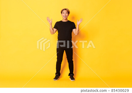 Full-length of surpirsed and excited, smiling blond asian guy raising hands up amazed, yellow background 85430029