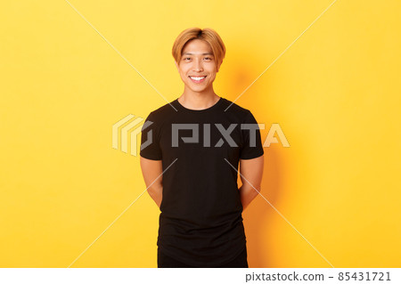 Portrait of friendly handsome asian guy with blond hair, smiling politely, holding hands behind back, standing yellow background 85431721