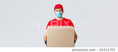 Groceries and packages delivery, covid-19, quarantine and shopping concept. Serious courier in red uniform, gloves and protective face mask, deliver package box to client house during coronavirus 85431763