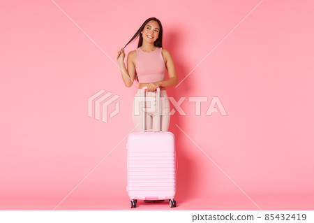 Travelling, holidays and vacation concept. Full-length of dreamy coquettish brunette asian girl in summer clothes thinking about her paradise resort, standing with suitcase over pink background 85432419
