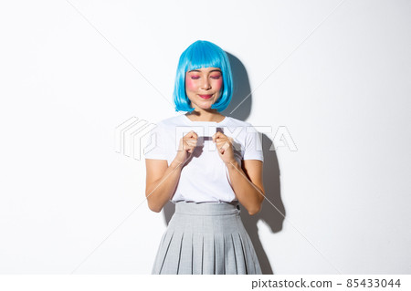 Portrait of beautiful asian girl in blue wig and halloween costume, daydreaming about shopping while holding credit card and smiling with closed eyes, standing over white background 85433044