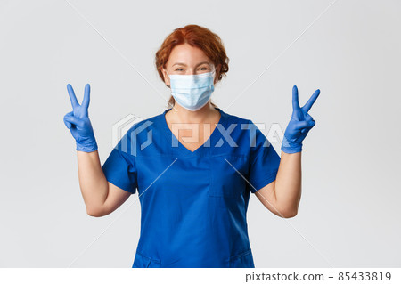 Medical workers, covid-19 pandemic, coronavirus concept. Optimistic smiling redhead female doctor, physician in medical mask and gloves showing peace gesture, staying positive during quarantine Medical workers, covid-19 pandemic, coronavirus concept. Optimistic smiling redhead female doctor, physician in medical mask and gloves showing peace gesture, staying positive during quarantine 85433819