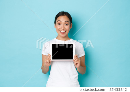 Excited and cheerful asian female, showing digital tablet screen and looking amazed, demonstrate her project or interesting link, advertising shopping site, standing light blue background 85433842