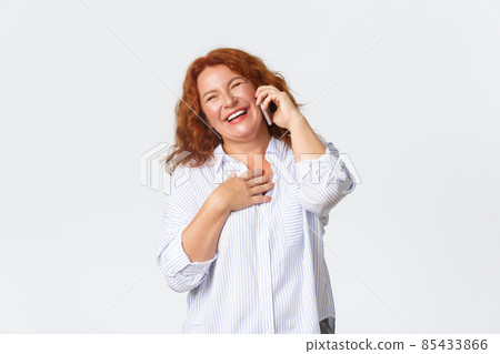 Portrait of cheerful, beautiful middle-aged redhead woman talking on phone, holding smartphone and laughing while having funny conversation, good cellphone receptions, cellular service 85433866