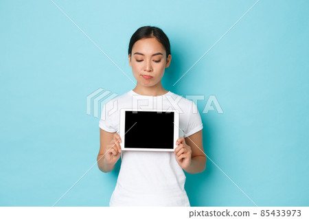 Skeptical and gloomy asian girl looking uneasy or doubtful, showing digital tablet screen, with perplexed expression, demonstrate something bad or complex, standing light blue background 85433973