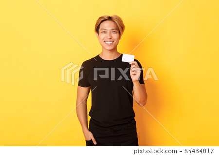 Cheerful handsome asian guy smiling pleased and showing credit card, standing yellow background Cheerful handsome asian guy smiling pleased and showing credit card, standing yellow background 85434017