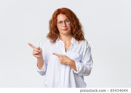 Skeptical and disappointed redhead middle-aged mother, woman in glasses scolding person, pointing fingers left at something bad, looking judgemental, standing white background Skeptical and disappointed redhead middle-aged mother, woman in glasses scolding person, pointing fingers left at something bad, looking judgemental, standing white background 85434044