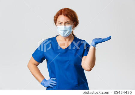 Medical workers, covid-19 pandemic, coronavirus concept. Confused skeptical female doctor, dentist in scrubs, face mask and gloves, shrugging, pointing right and frowning disappointed 85434450