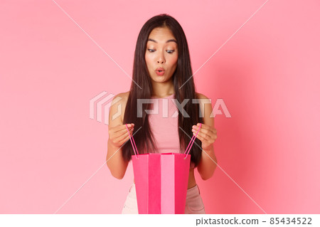 Beauty, fashion and lifestyle concept. Close-up of excited and amazed, surprised girl open up present, looking in shopping bag with impressed and wondered expression, pink background Beauty, fashion and lifestyle concept. Close-up of excited and amazed, surprised girl open up present, looking in shopping bag with impressed and wondered expression, pink background 85434522
