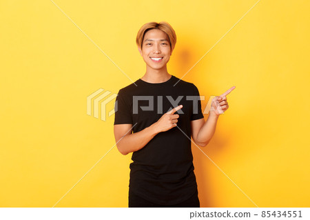 Portrait of satisfied smiling blond guy, asian student pointing fingers right, showing way, standing yellow background Portrait of satisfied smiling blond guy, asian student pointing fingers right, showing way, standing yellow background 85434551