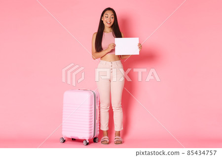 Travelling, holidays and vacation concept. Full-length of coquettish trendy brunette asian girl waiting for someone to pick her up airport, standing with sign and suitcase over pink background 85434757