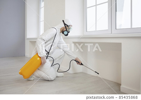Pest control exterminator getting rid of insects inside infested house. Man in protective mask and suit spraying insecticide from yellow bottle on floor and walls for safe living environment at home 85435868
