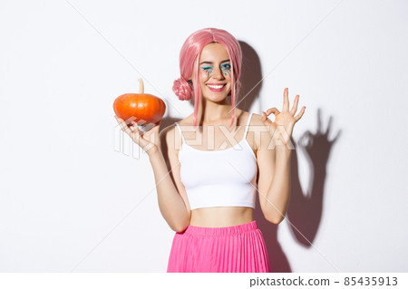 Image of happy girl celebrating halloween, wearing pink wig, holding pumpkin and showing okay sign, standing over white background 85435913