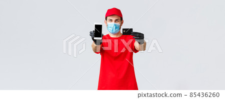 Contactless delivery, payment and online shopping during covid-19, self-quarantine. Friendly courier in red uniform, face mask and gloves, showing smartphone screen and credit card, order internet 85436260