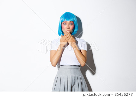 Portrait of surprised beautiful asian girl in glamour blue wig, looking amazed, holding hands near lips thrilled, standing over white background 85436277