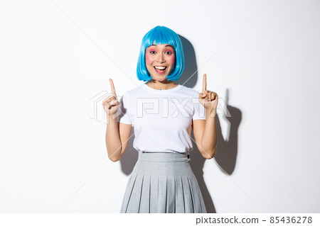Joyful good-looking asian girl in blue wig, celebrating holiday, dressed for party or halloween, pointing fingers up and smiling happy, standing over white background 85436278
