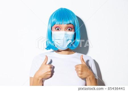 Concept of halloween celebration and coronavirus. Close-up of excited asian girl in medical mask and blue wig showing thumbs-up and approve something good 85436441
