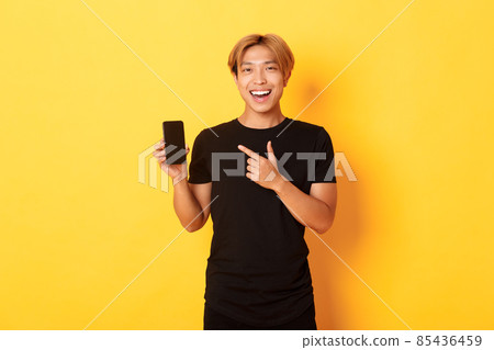Portrait of satisfied smiling asian guy with blond hair, looking amused and pointing finger at smartphone screen, standing yellow background 85436459