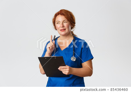 Medicine, healthcare and coronavirus concept. Thoughtful redhead female doctor, redhead physician in blue scrubs looking intrigued with patient case, shaking pen and holding clipboard 85436460