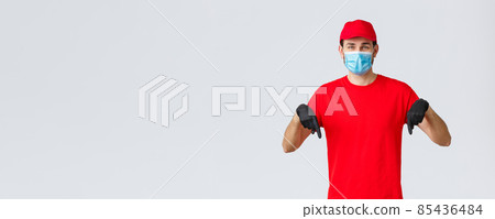 Covid-19, self-quarantine, online shopping and shipping concept. Smiling friendly delivery guy red uniform, cap and t-shirt, wear medical mask with gloves, pointing down, showing promo or client info Covid-19, self-quarantine, online shopping and shipping concept. Smiling friendly delivery guy red uniform, cap and t-shirt, wear medical mask with gloves, pointing down, showing promo or client info 85436484