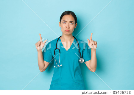 Covid-19, social distancing and coronavirus pandemic concept. Disappointed gloomy asian female doctor, nurse complaining over something bad, sulking and pointing fingers up Covid-19, social distancing and coronavirus pandemic concept. Disappointed gloomy asian female doctor, nurse complaining over something bad, sulking and pointing fingers up 85436738