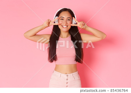Portrait of beautiful smiling asian girl in wireless headphones, listening music, enjoying sound quality, showing peace or kawaii gestures voer eyes and grinning happy over pink background 85436764