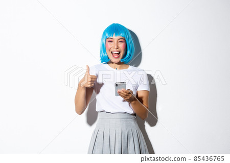 Portrait of pretty asian woman in blue wig laughing happy, showing thumbs-up, holding mobile phone Portrait of pretty asian woman in blue wig laughing happy, showing thumbs-up, holding mobile phone 85436765