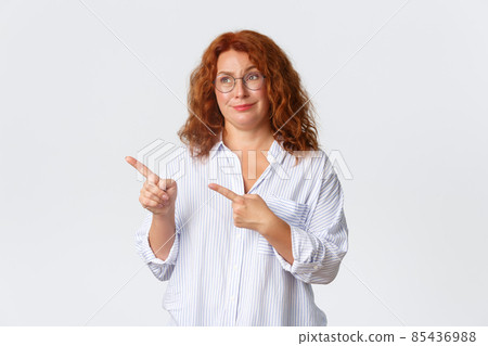 Skeptical and unamused, smirking redhead middle-aged woman looking and pointing at something average, feeling reluctant about promo banner, dont like product, standing white background Skeptical and unamused, smirking redhead middle-aged woman looking and pointing at something average, feeling reluctant about promo banner, dont like product, standing white background 85436988