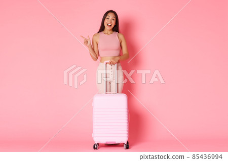 Travelling, holidays and vacation concept. Full-length of amused beautiful asian girl in summer clothes, explore countries, enjoying summer abroad, pointing left, stand with suitcase, pink background 85436994