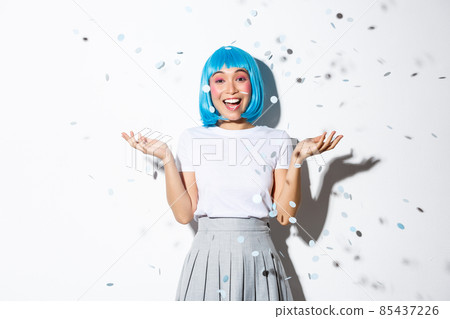 Portrait of happy asian girl in blue wig celebrating halloween, throwing confetti in the air, standing over white background Portrait of happy asian girl in blue wig celebrating halloween, throwing confetti in the air, standing over white background 85437226
