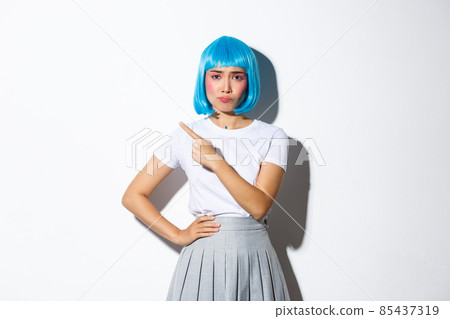 Image of skeptical and unamused asian girl in blue party wig, grimacing upset and pointing finger at upper left corner, standing against white background 85437319