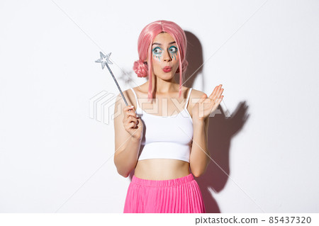 Image of amused cute girl with pink wig and bright makeup, dressed up as fairy for halloween party, holding magic wand and looking excited, standing over white background 85437320