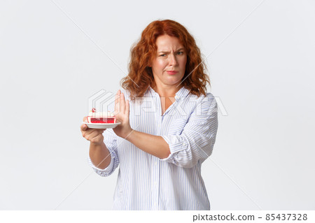 Holidays, emotions and lifestyle concept. Displeased frowning and grimacing redhead middle-aged woman holding bad cake, showing refusal gesture, rejecting dessert, white background 85437328