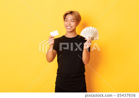 Portrait of satisfied smiling, handsome asian guy looking pleased at credit card while also holding money, standing yellow background Portrait of satisfied smiling, handsome asian guy looking pleased at credit card while also holding money, standing yellow background 85437656