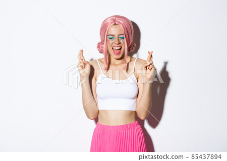 Image of optimistic party girl in pink wig, with halloween makeup, close eyes and screaming excited while cross fingers to make a wish, standing over white background 85437894