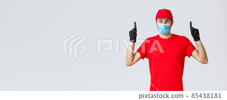 Covid-19, self-quarantine, online shopping and shipping concept. Amused happy delivery guy advertise courier company service during coronavirus lockdown, pointing fingers up, wear medical mask 85438181