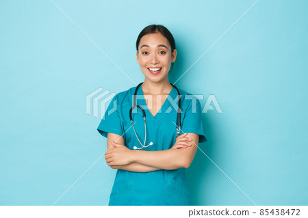 Covid-19, social distancing and coronavirus pandemic concept. Cheerful smiling asian female doctor, physician or nurse in scrubs cross arms and discuss something, standing light blue background 85438472