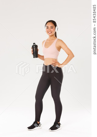 Full length of slim and healthy asian girl doing fitness, female athelte workout in gym, listening music in wireless headphones, drinking water from bottle during active training, white background 85438485