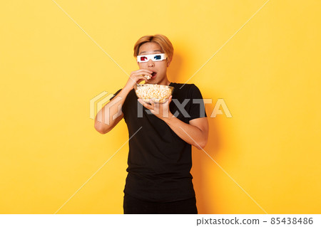 Impressed and thrilled asian guy attend cinema, watching movie or tv series in 3d glasses, eating popcorn and looking shocked, standing yellow background 85438486