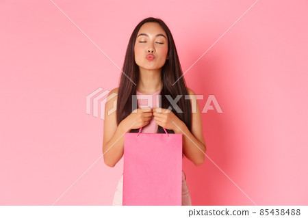 Beauty, fashion and lifestyle concept. Portrait of glamour flirty asian girlfriend holding shopping bag with clothes boyfriend bought, leaning for kiss and close eyes sensual, pink background 85438488