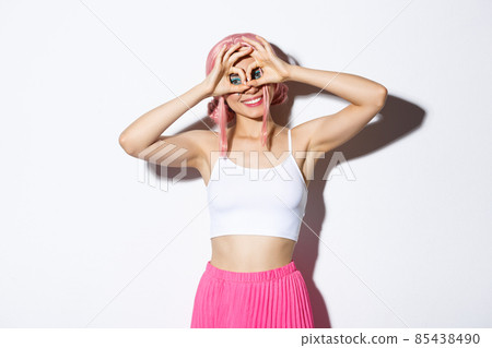 Image of funny cute girl celebrating halloween, wearing pink anime wig, making finger glasses and grimacing, standing over white background 85438490