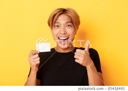 Close-up of happy and satisfied asian handsome guy, showing credit card and thumbs-up in approval, smiling amazed, standing yellow background 85438491