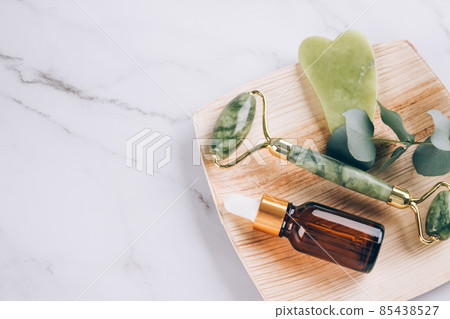 Jade gua sha facial roller with beauty serum oil on white marble table background with copy space. Facial massage kit for lifting massage therapy made from natural stones 85438527