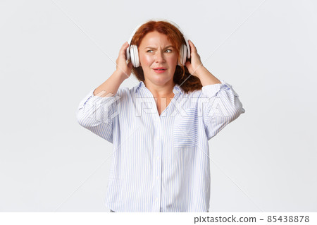 Emotions, lifestyle and leisure concept. Confused and displeased redhead middle-aged female using headphones, cant understand what playing in earphones, standing white background disappointed Emotions, lifestyle and leisure concept. Confused and displeased redhead middle-aged female using headphones, cant understand what playing in earphones, standing white background disappointed 85438878