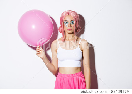 Image of young stylish girl holding pink balloon, wearing party wig and celebrating holiday, standing over white background Image of young stylish girl holding pink balloon, wearing party wig and celebrating holiday, standing over white background 85439179