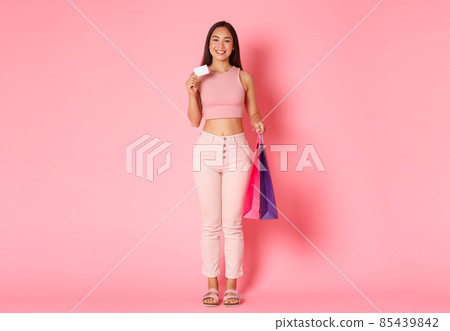 Full length of trendy glamour asian girl in summer clothes enjoying vacation, shopping with her credit card in foreign countries, holding bags with purchased items, pink background 85439842