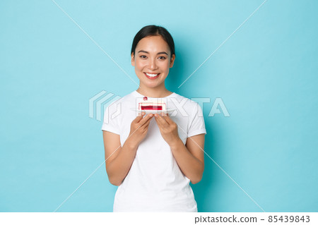 Holidays, lifestyle and celebration concept. Smiling beautiful slim asian girl in white t-shirt, showing piece of cake and looking upbeat, eating delicious dessert, standing light blue background 85439843