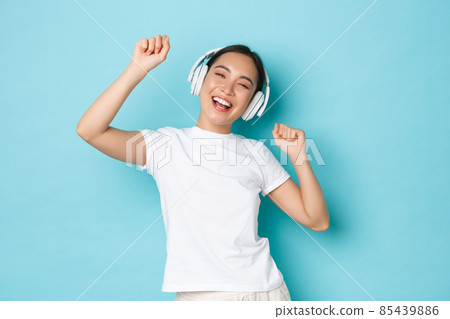 Shopping, money and finance concept. Upbeat asian girl in white casual t-shirt, singing karaoke, playing music app, listening music in wireless earphones and enjoying perfect sound 85439886