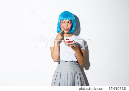 Coquettish beautiful asian girl in blue short wig, looking thoughtful and cunning at upper left corner, holding piece of cake, standing over white background 85439888