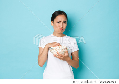 Lifestyle, leisure and emotions concept. Skeptical and displeased asian cute girl watching movie and eating popcorn, grimacing from cringe content, looking suspicious, standing light blue background 85439894
